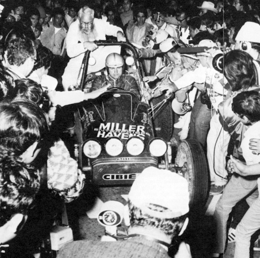 Surrounded by swarming press and excited locals, Drino Miller pulls his "Dune Buggies and Hot VW's" -sponsored racer into victory lane as the overall winner of the 1970 NORRA Mexican 1000. The innovative VW-powered single-seater was built and designed by Miller and co-driven by inaugural Mexican 1000 champion Vic Wilson. Note NORRA official Elmer Waring (in white) hitching a ride. 